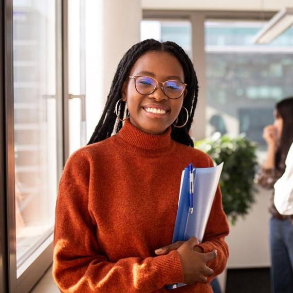 A person wearing an orange sweater holds a blue folder while standing by a window in an office setting